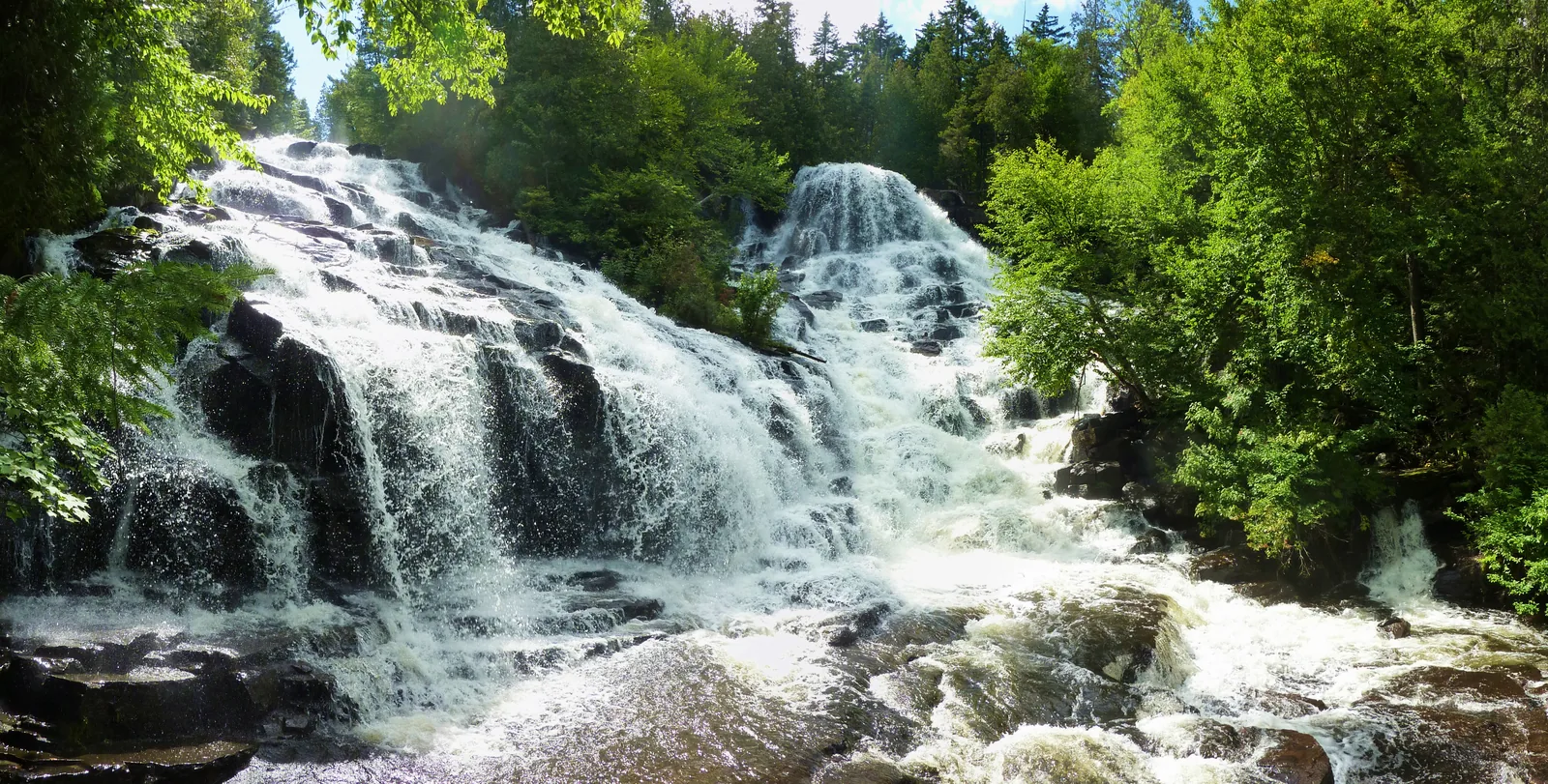 Ein breiter Wasserfall stürzt über mehrere Stufen dunklen Felsens, ringsum dichter grüner Laubwald