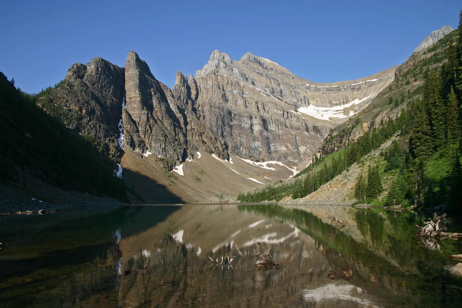 Lake Agnes im Banff-Nationalpark spiegelt die Felswände des Mount Whyte, rechts bewaldete Hänge und Schneefelder im Kar.