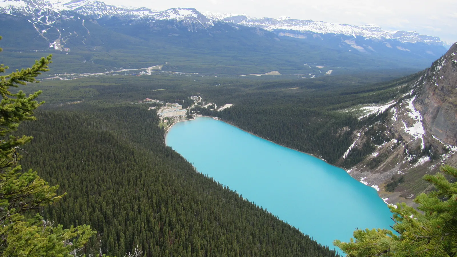 Blick von oben auf den türkisfarbenen Lake Louise zwischen Wäldern, am Ende das Hotel