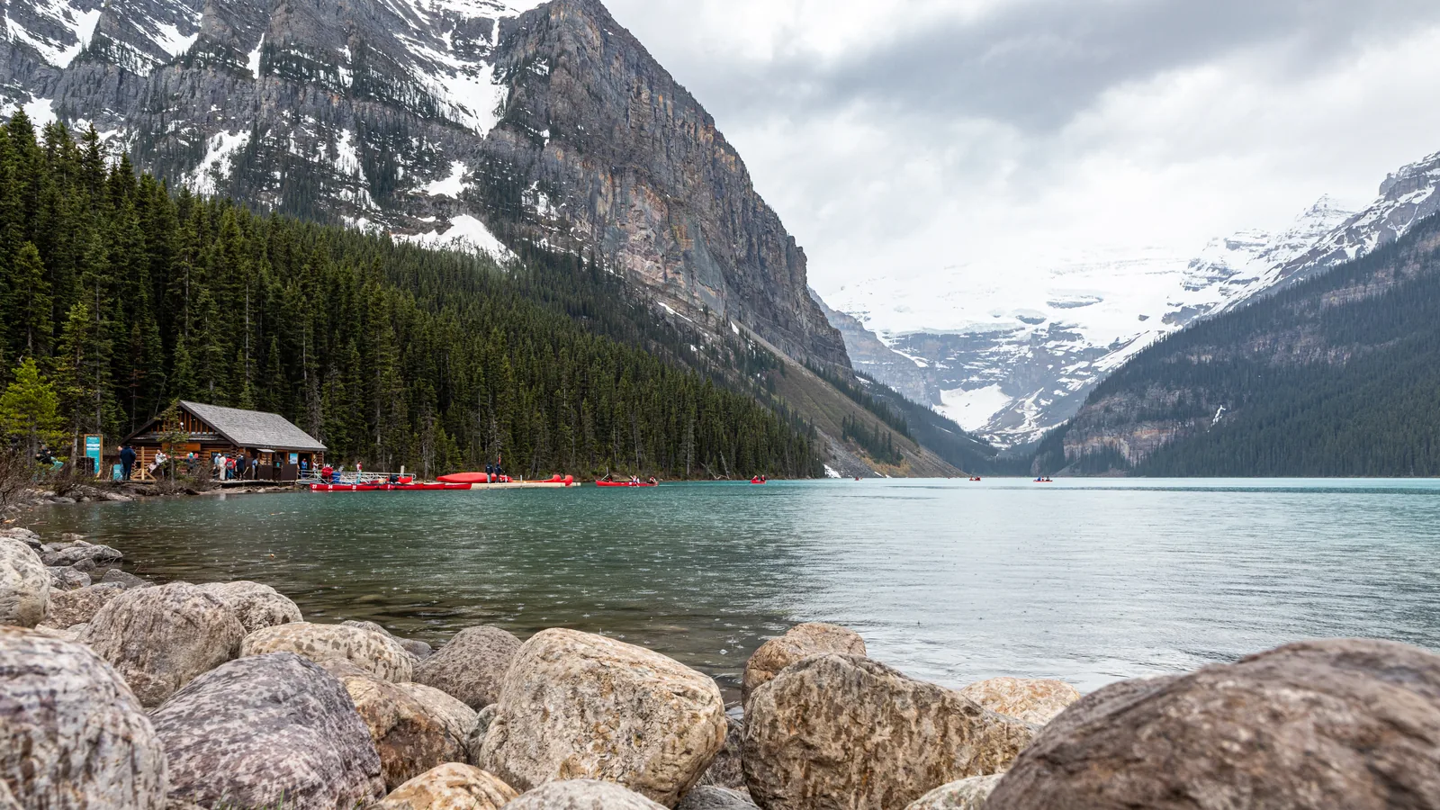 Lake Louise vom Ostufer: Felsbrocken im Vordergrund, Bootshaus und Kanus am Ufer, hinten der Gletscher