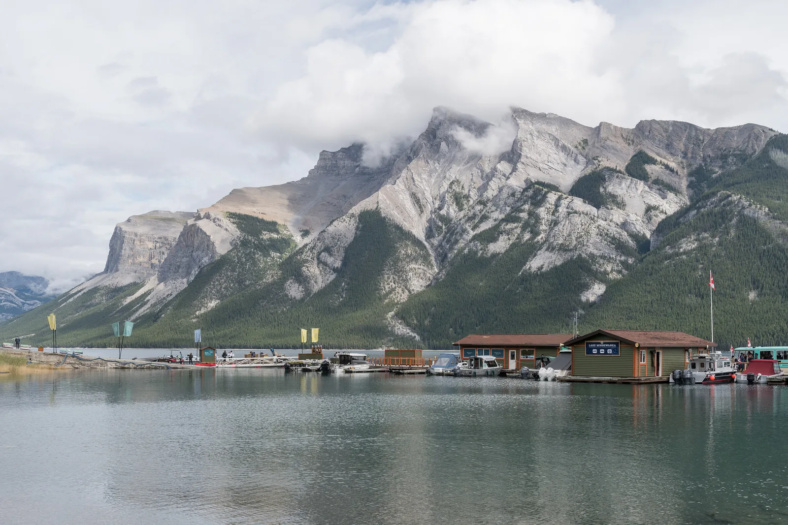 Bootssteg am Lake Minnewanka im Banff-Nationalpark mit Bootshaus und Ausflugsbooten, dahinter der Mount Inglismaldie in Wolken.