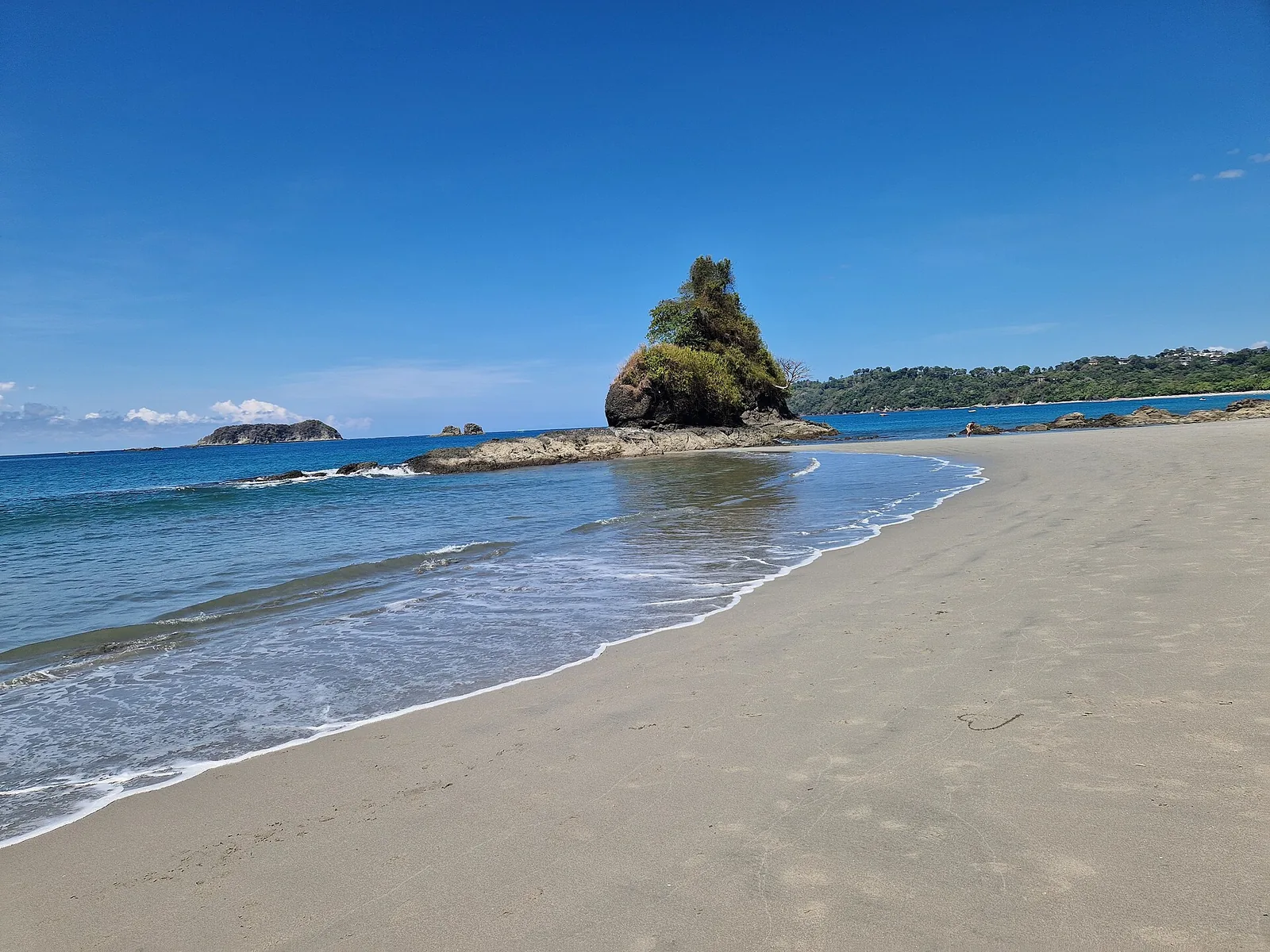 Heller Sandstrand im Manuel-Antonio-Nationalpark mit einer bewaldeten Felsinsel im türkisfarbenen Pazifik unter blauem Himmel