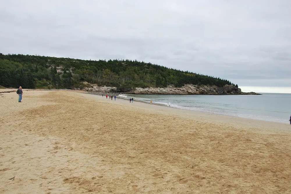 Sand Beach bei der Gebührenstation: breiter Sandstrand zwischen bewaldeter Landspitze und Meer, einzelne Spaziergänger
