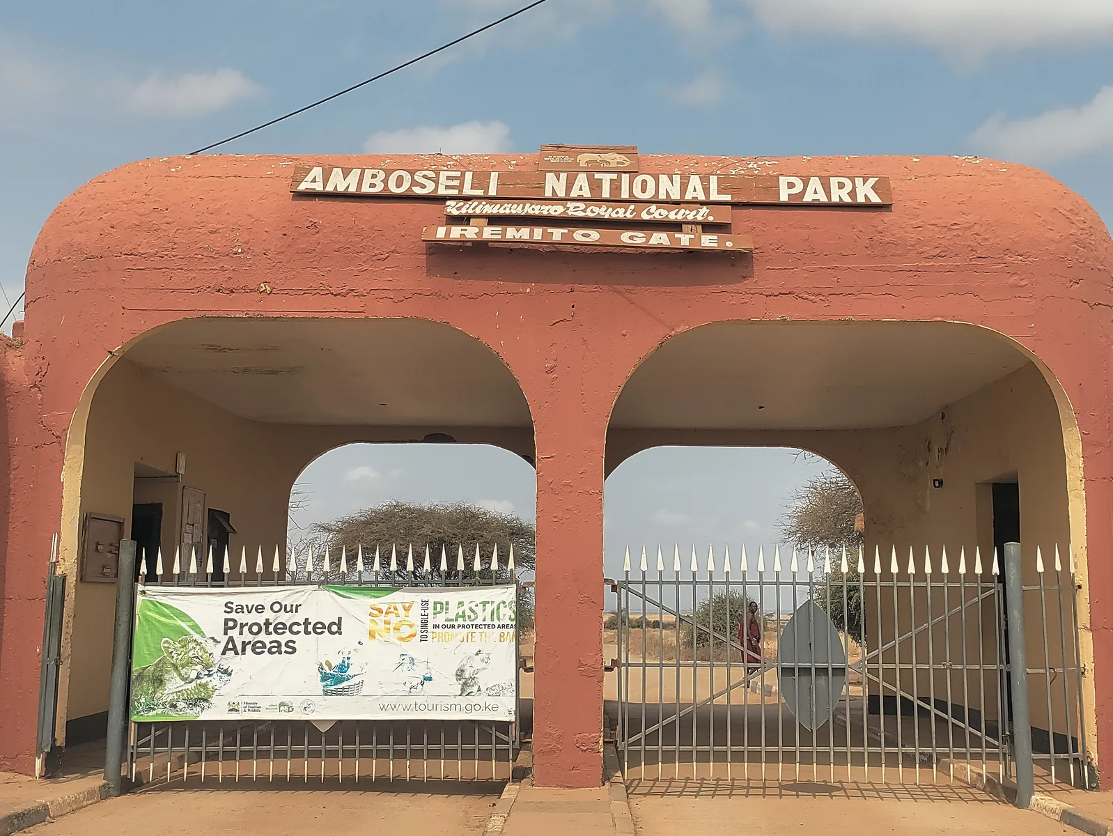 Iremito Gate des Amboseli-Nationalparks: rotbraunes Torgebäude mit zwei Bögen und der Aufschrift Amboseli National Park, davor ein geschlossenes Gittertor