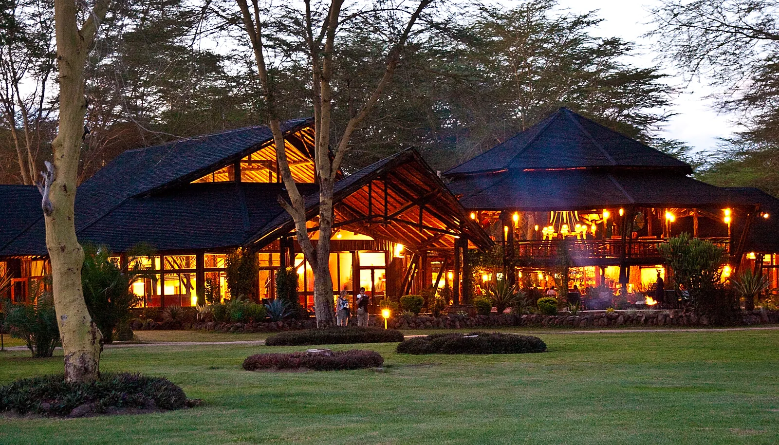Das beleuchtete Hauptgebäude der Ol Tukai Lodge im Amboseli-Nationalpark am Abend, mit hohen Spitzdächern zwischen Akazien