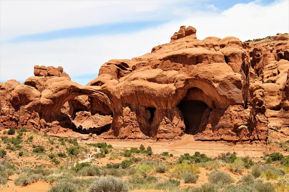Double Arch: zwei ineinander verschränkte Sandsteinbögen in einer roten Felswand, davor niedrige Wüstenvegetation
