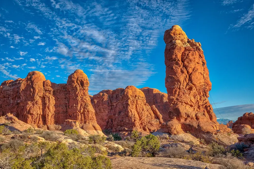 Garden of Eden: rote Sandsteinsäulen und Felstürme unter blauem Schleierwolkenhimmel, davor niedrige Wüstenvegetation