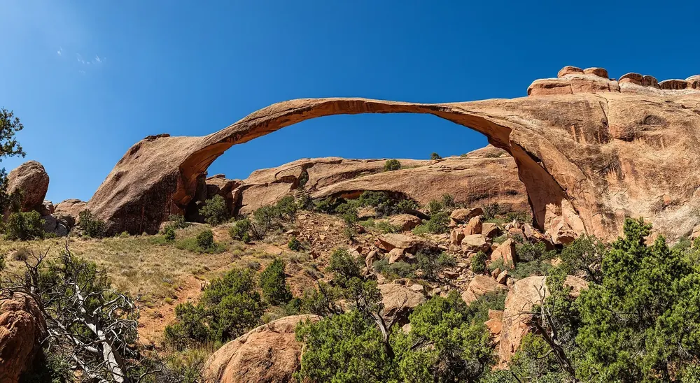 Landscape Arch: extrem langer, dünner Sandsteinbogen über Felsblöcken und grünem Wacholder unter tiefblauem Himmel