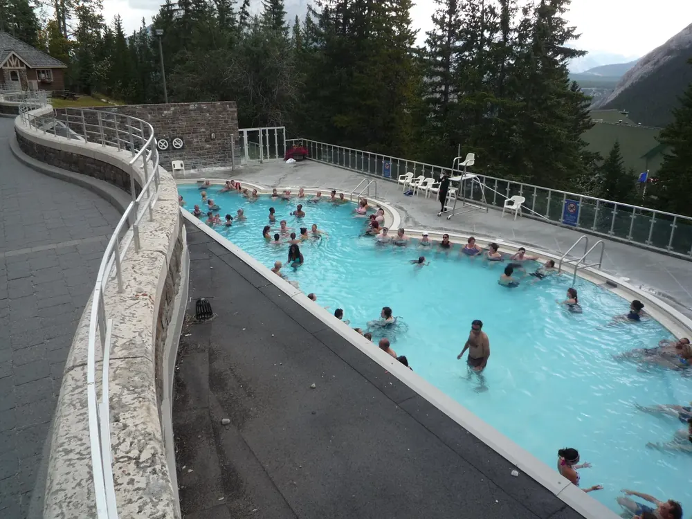 Außenbecken der Banff Upper Hot Springs mit Badegästen im türkisfarbenen Thermalwasser, dahinter Nadelwald am Hang