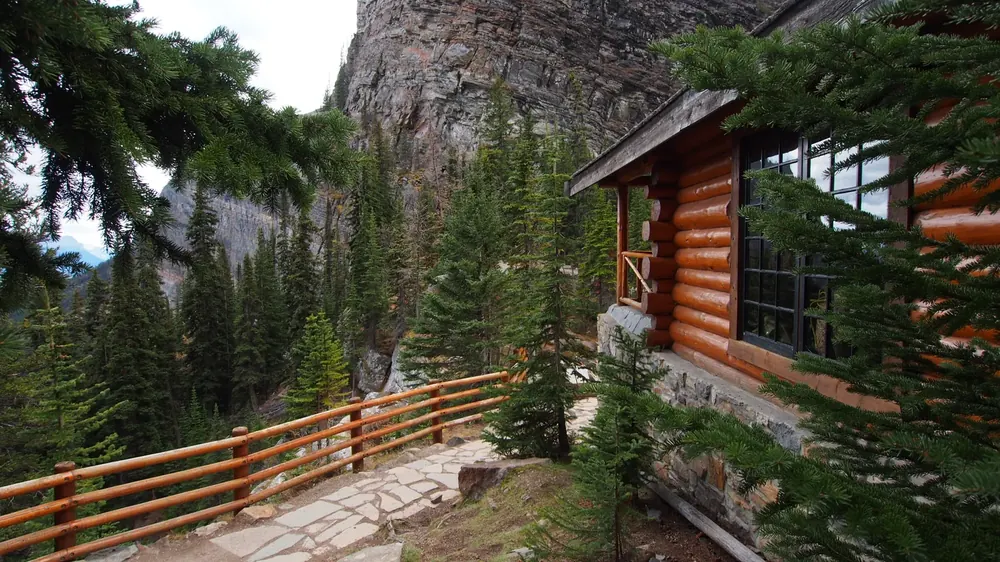 Holzblockhaus des Lake Agnes Tea House mit Geländer und Steinterrasse zwischen Nadelbäumen unter einer Felswand