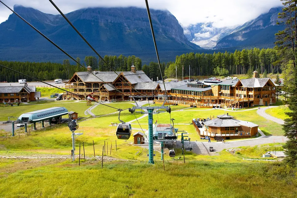 Talstation des Skigebiets Lake Louise im Sommer: Gondelbahn, Holzlodges und grüne Wiesen vor bewaldeten Bergen