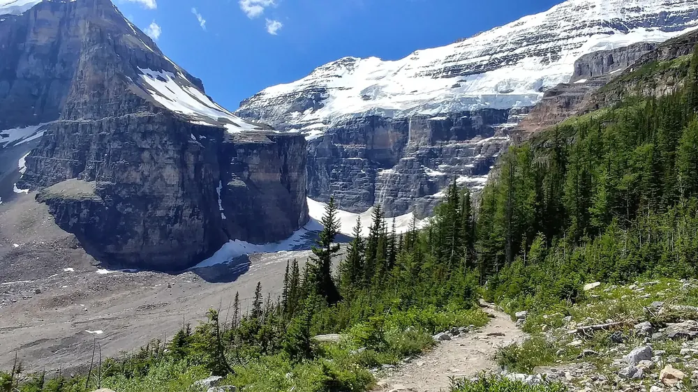 Weg durch die Plain of Six Glaciers: Geröllhänge, Nadelwald und vergletscherte Felswände unter blauem Himmel