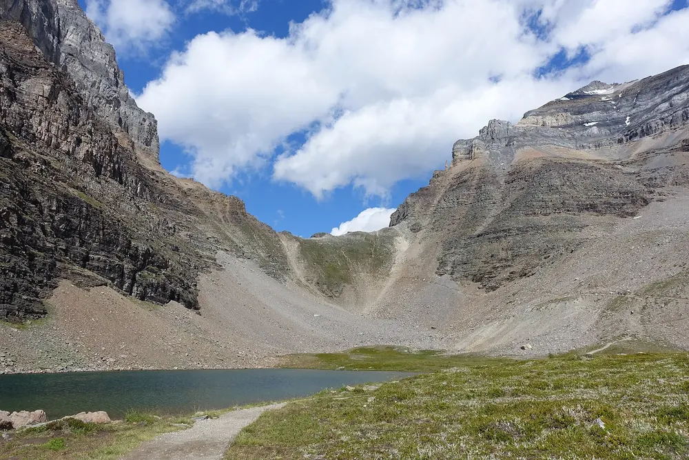 Sentinel Pass: Geröllsattel zwischen steilen Felswänden, davor ein kleiner Bergsee und ein Wanderweg über Almwiesen