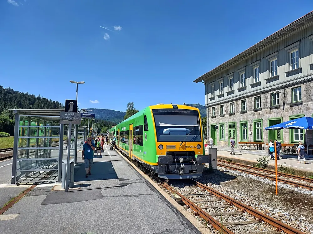 Triebwagen der Waldbahn im Grenzbahnhof Bayerisch Eisenstein, rechts das historische Bahnhofsgebäude, dahinter bewaldete Berge