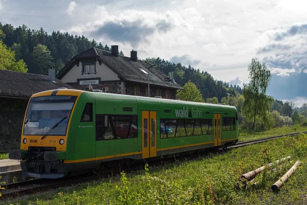 Grün-gelber Triebwagen der Waldbahn im Bahnhof Grafenau, dahinter das Bahnhofsgebäude und bewaldete Hügel