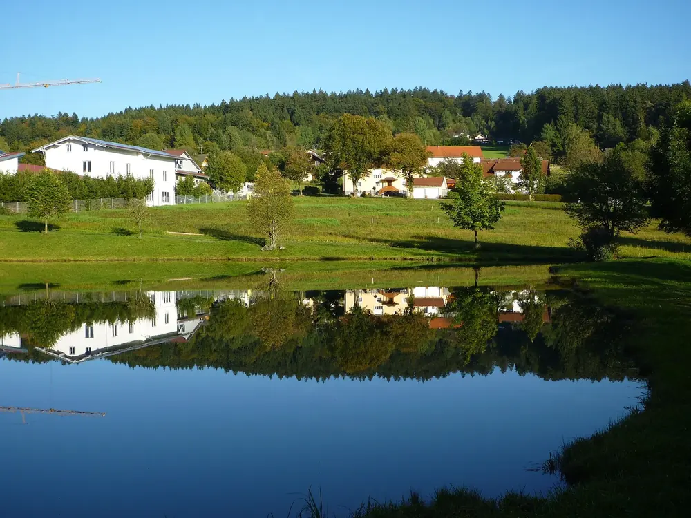 Badesee in Neuschönau mit Spiegelung, dahinter Wiesen, Häuser des Ortes und bewaldete Hügel