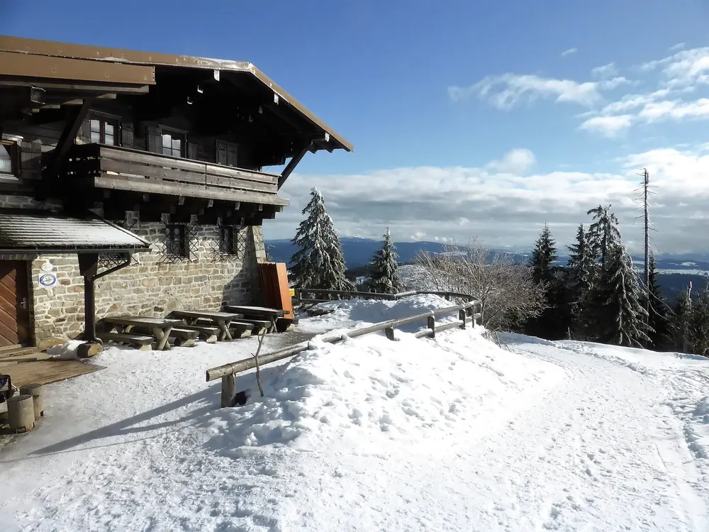 Lusen-Schutzhaus im Winter: Berghütte aus Stein und Holz, davor verschneite Terrasse, dahinter verschneite Fichten und Fernsicht