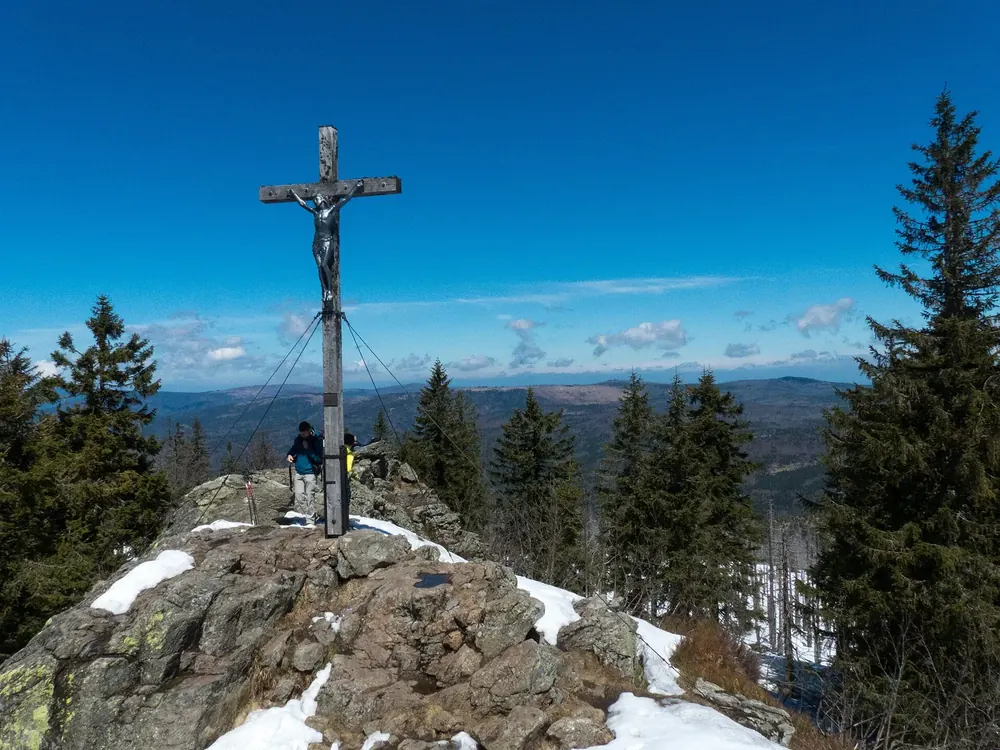 Gipfelkreuz auf dem felsigen Großen Rachel, ringsum Fichten und Schneereste, Weitblick über den Bayerischen Wald