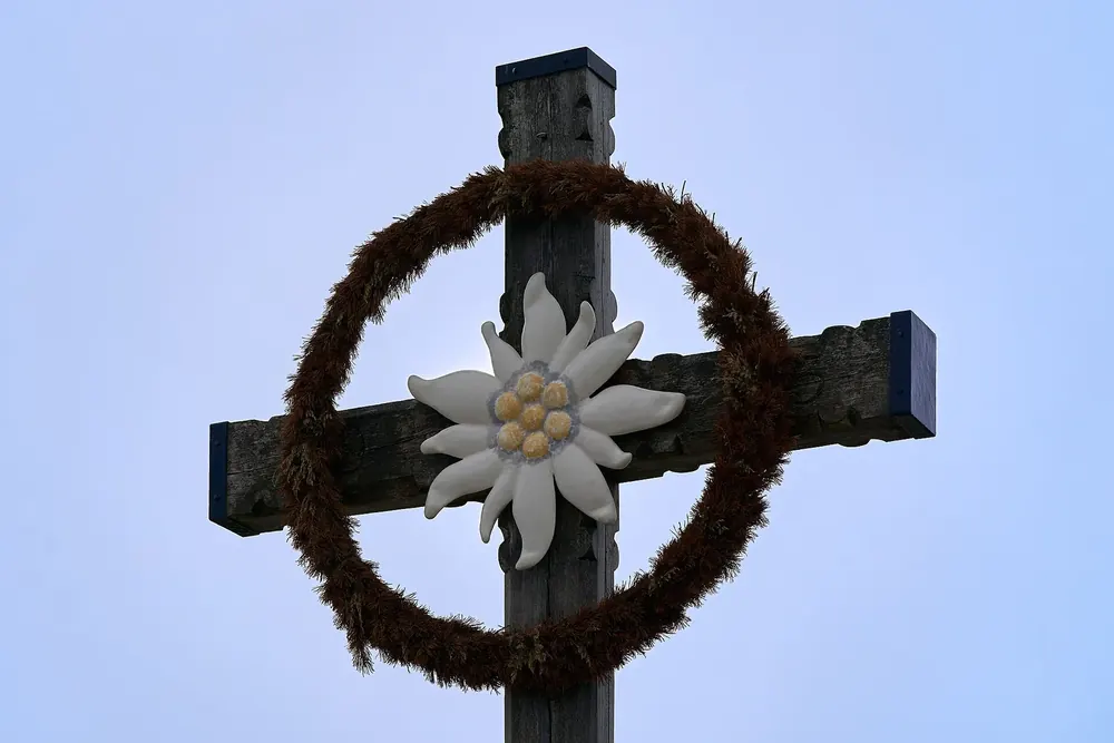 Hölzernes Gipfelkreuz auf dem Kehlstein mit geflochtenem Kranz und großem Edelweiß vor blauem Himmel