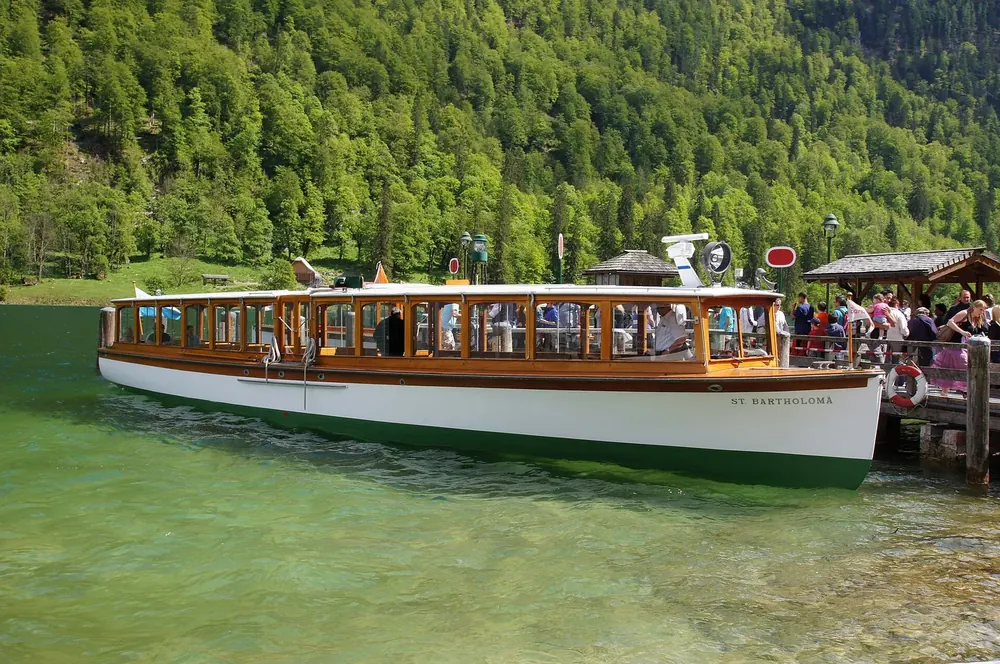 Ausflugsboot an der Anlegestelle St. Bartholomä im grünen Wasser des Königssees, dahinter bewaldeter Steilhang