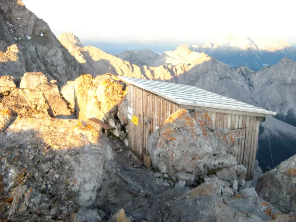 Kleine hölzerne Schutzhütte auf dem felsigen Hocheck des Watzmann, dahinter Bergketten im Abendlicht