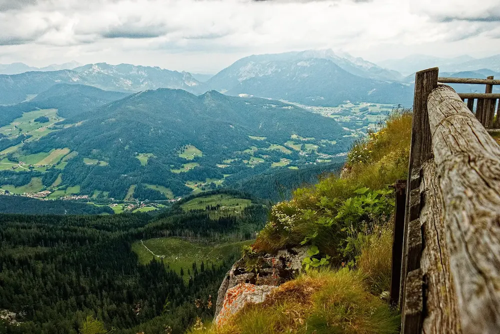 Blick vom Watzmannhaus über das Holzgeländer der Terrasse hinab ins Berchtesgadener Tal und auf ferne Bergketten