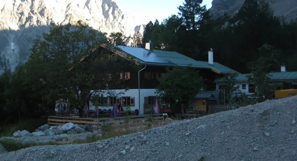 Wimbachgrieshütte am Rand des hellen Schotterfelds im Wimbachgries, dahinter Nadelbäume und Felswände im Abendlicht