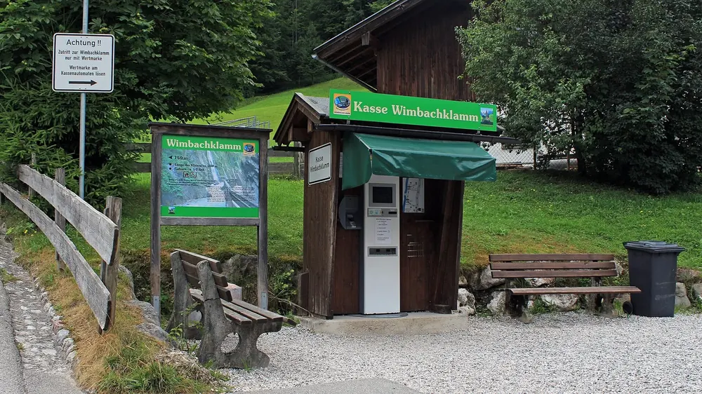 Kassenhäuschen der Wimbachklamm nahe dem Parkplatz Wimbachbrücke, daneben Infotafel, Bänke und Laubwald