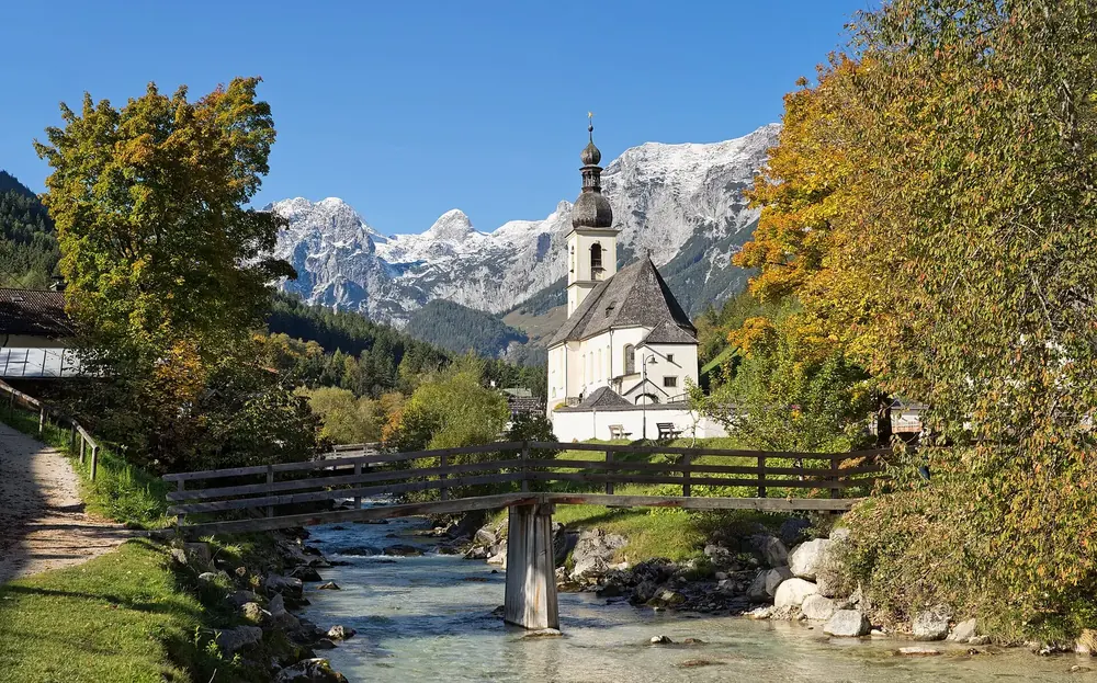 Pfarrkirche St. Sebastian in Ramsau mit Holzbrücke über den Bach, dahinter Herbstlaub und verschneite Gipfel