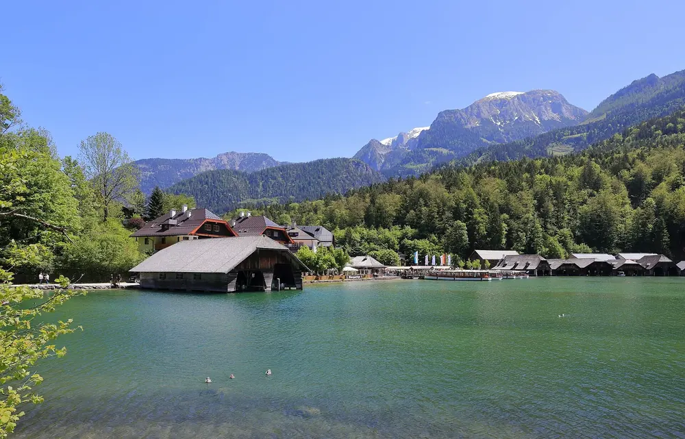 Königssee bei Schönau: Bootshäuser am grünen Ufer, dahinter bewaldete Hänge und Gipfel der Berchtesgadener Alpen