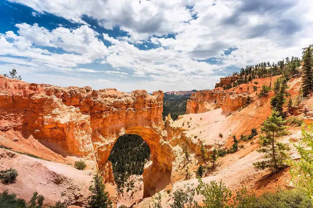 Natural Bridge: rotorangefarbener Felsbogen über bewaldetem Grund, umgeben von Erosionshängen unter Quellwolken