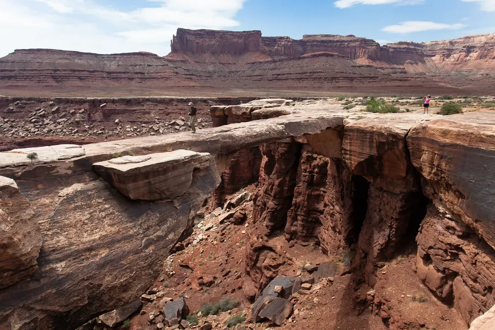 Musselman Arch: flacher Sandsteinbogen über einer tiefen Felsspalte, dahinter rote Tafelberge am Horizont