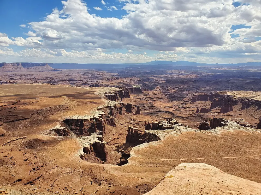 Blick vom White Rim Overlook auf verzweigte Canyons mit hellem Felsband und ferne Tafelberge unter Wolken