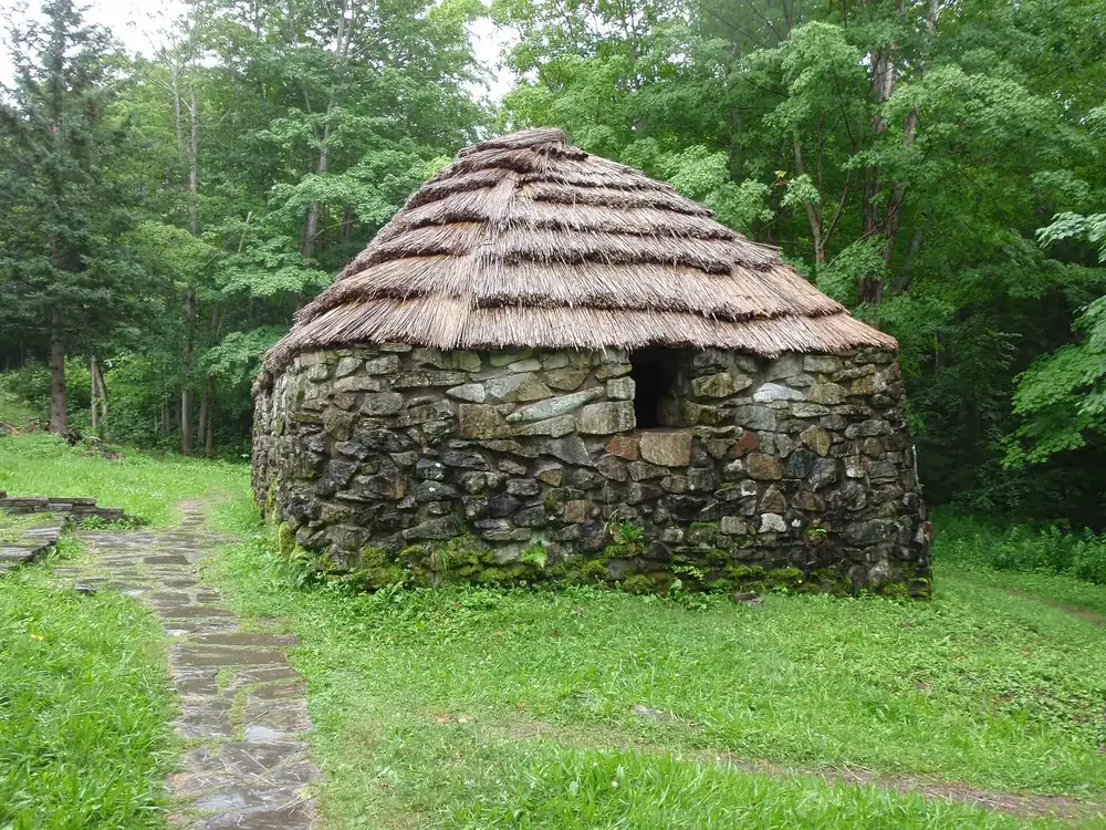Lone Shieling: nachgebaute schottische Hirtenhütte aus Feldsteinen mit Strohdach, umgeben von Laubwald