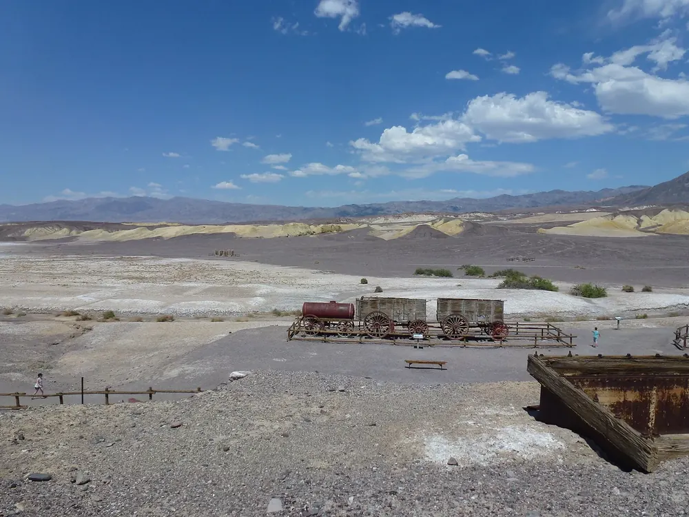 Harmony Borax Works: Reste der Boraxanlage mit historischen Wagen auf karger Ebene, dahinter Hügel und Berge