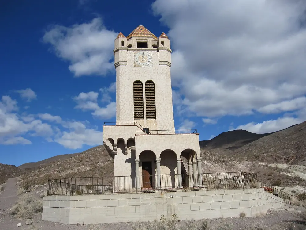Scotty’s Castle: heller Turm mit Uhr und Arkaden, dahinter karge Wüstenhügel unter blauem Himmel