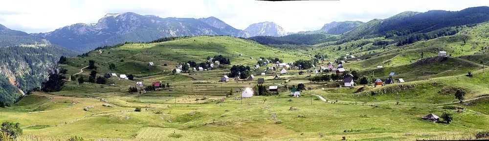Panorama des Dorfes Nedajno auf der grünen Hochebene, ringsum Wiesen, dahinter Berge des Durmitor