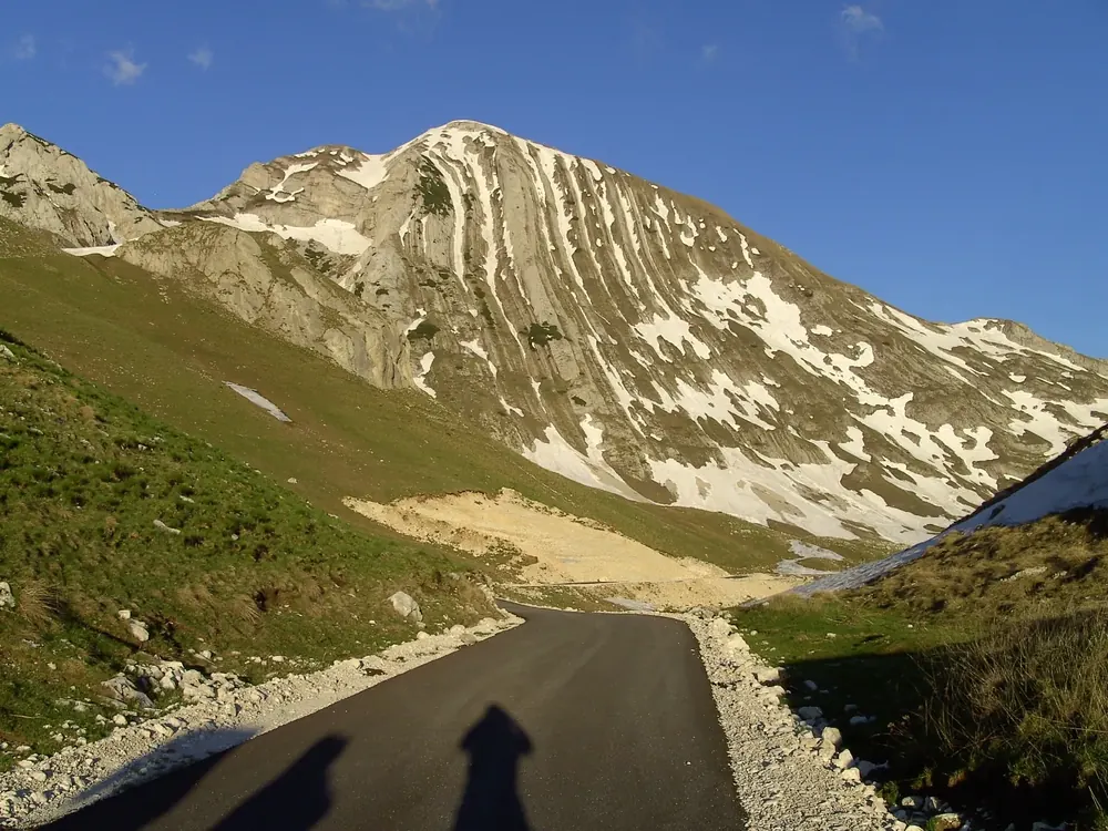 Gipfel des Prutaš mit auffällig gefalteten Gesteinsschichten und Schneeresten, davor eine Bergstraße im Hochtal