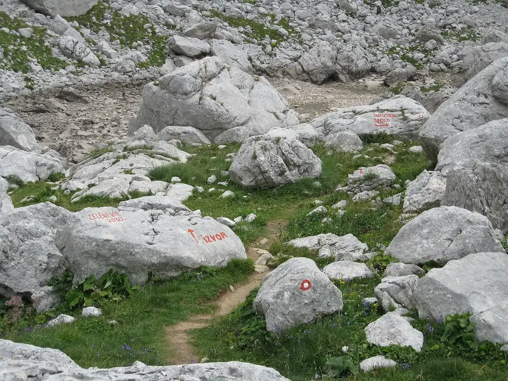 Ausgetrocknetes Becken des Zeleni Vir: helle Kalkblöcke und Grasflecken, rote Wegmarkierungen auf den Steinen