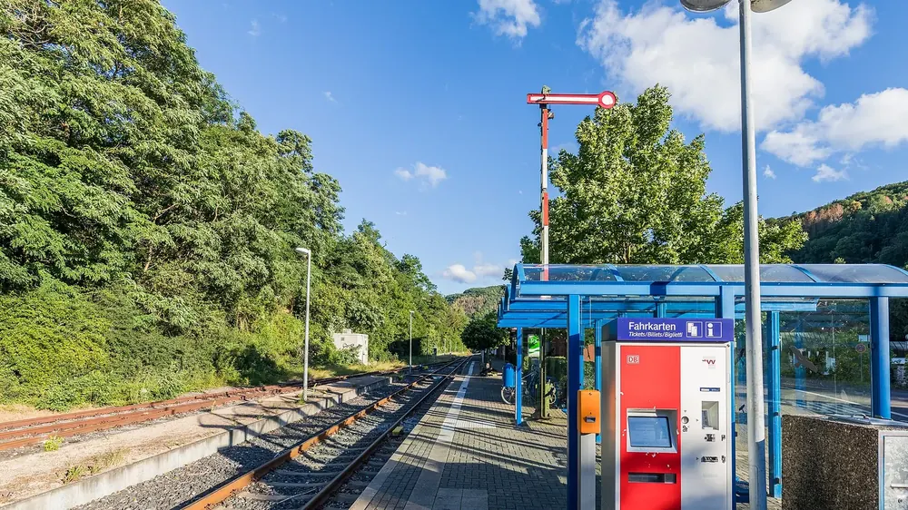 Bahnhof Heimbach: Bahnsteig mit Fahrkartenautomat und gläsernem Wartehäuschen, dahinter bewaldete Hänge