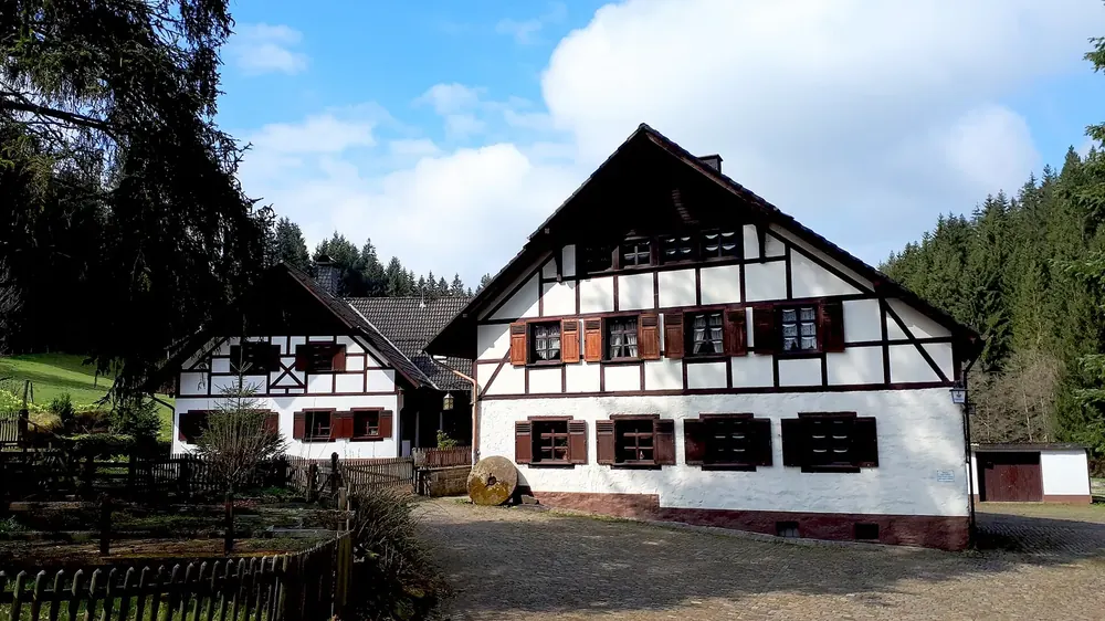 Ehemalige Höfener Mühle in Monschau-Höfen: weißes Fachwerkhaus mit roten Fensterläden am Waldrand
