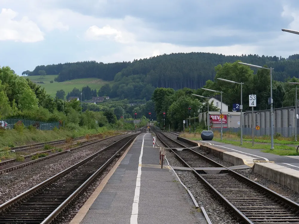 Bahnhof Kall: Bahnsteige und Gleise der Eifelstrecke, dahinter bewaldete Hügel unter bewölktem Himmel