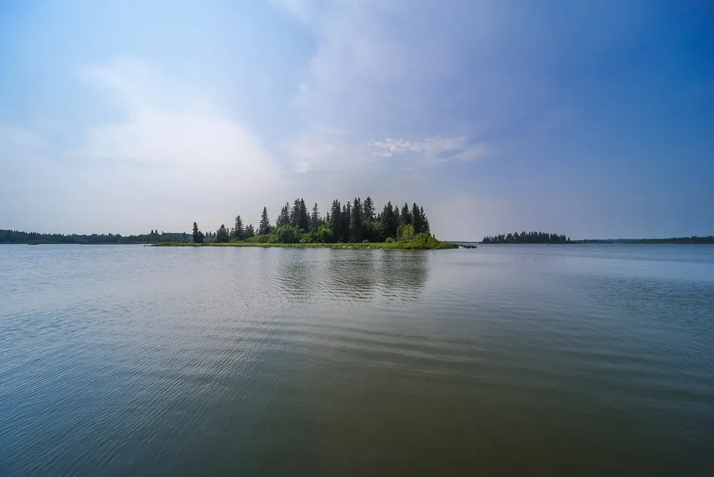 Astotin Lake beim Campingplatz: stiller See mit bewaldeter Insel unter hellem Himmel