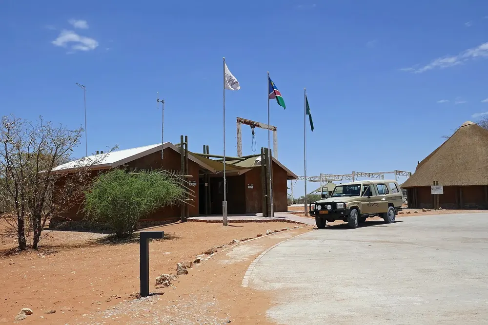 Olifantsrus Camp: Empfangsgebäude mit Fahnen, Reetdachhütte und Geländewagen unter blauem Himmel