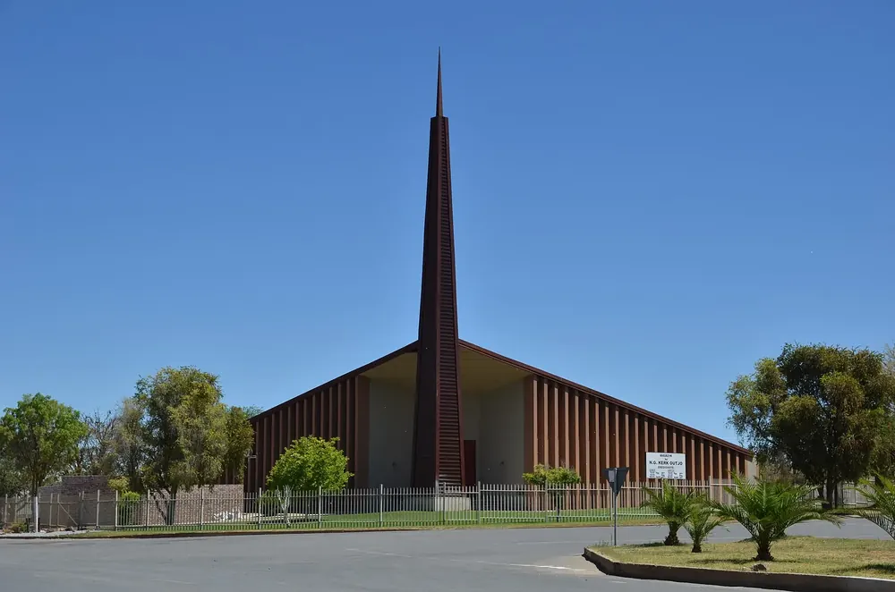 Kirche in Outjo mit spitzem, hohem Turm und steilem Dach, davor Bäume und Palmen unter blauem Himmel