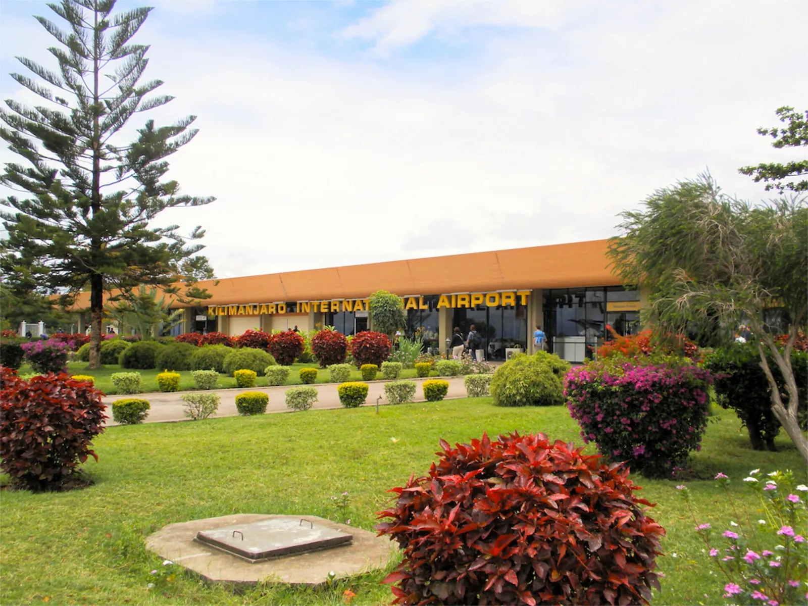 Terminal des Kilimanjaro International Airport mit Schriftzug hinter einem Garten mit roten Sträuchern und Blumenbeeten