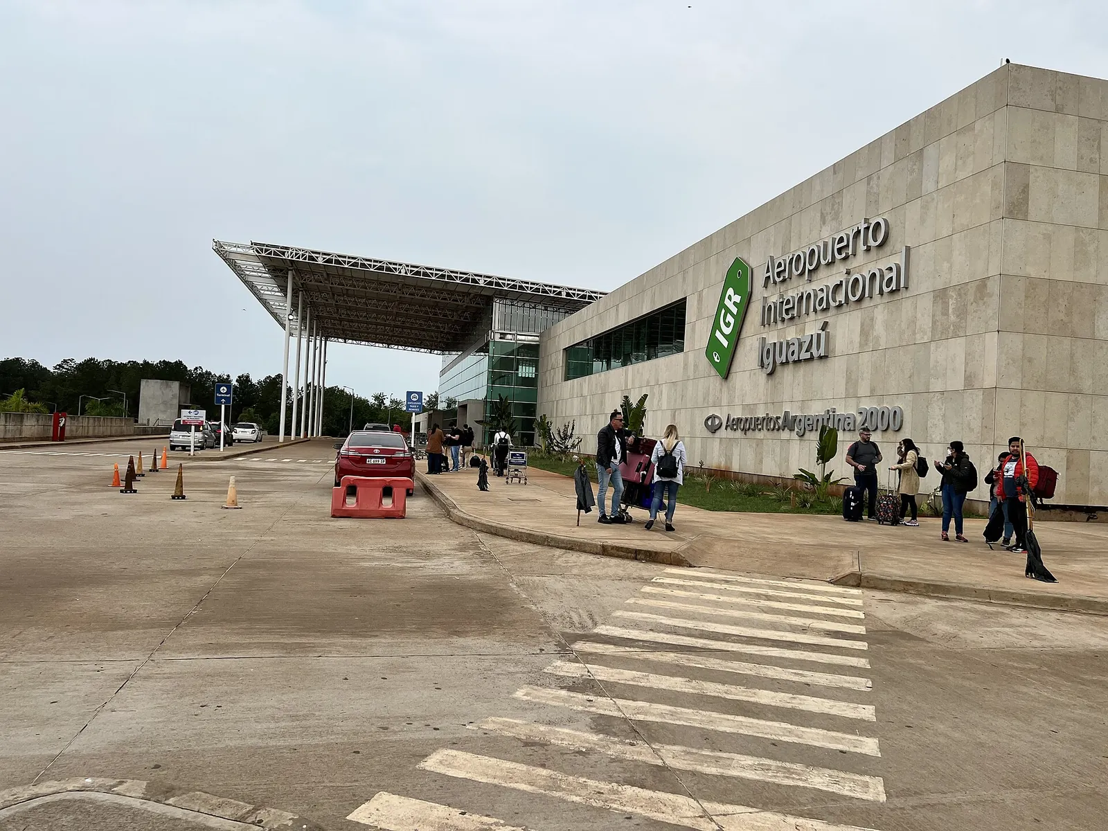 Flughafen Puerto Iguazú: Terminal mit Natursteinfassade und dem Schriftzug „Aeropuerto Internacional Iguazú“, davor ein Zebrastreifen und Reisende mit Gepäck