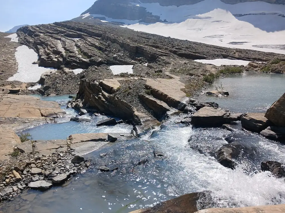 Schmelzwasser des Grinnell-Gletschers stürzt über geschichtete Kalkfelsen, dahinter Schneefelder und Eis