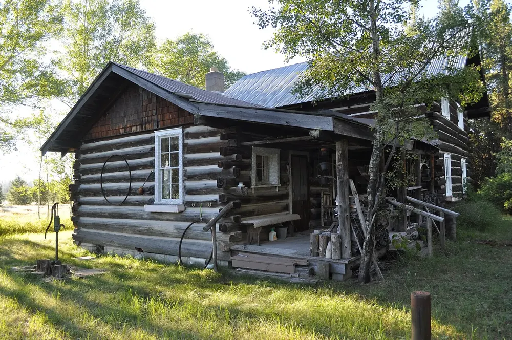 Vance Lodge in Polebridge: historisches Blockhaus mit Veranda und Blechdach auf einer Wiese zwischen Laubbäumen