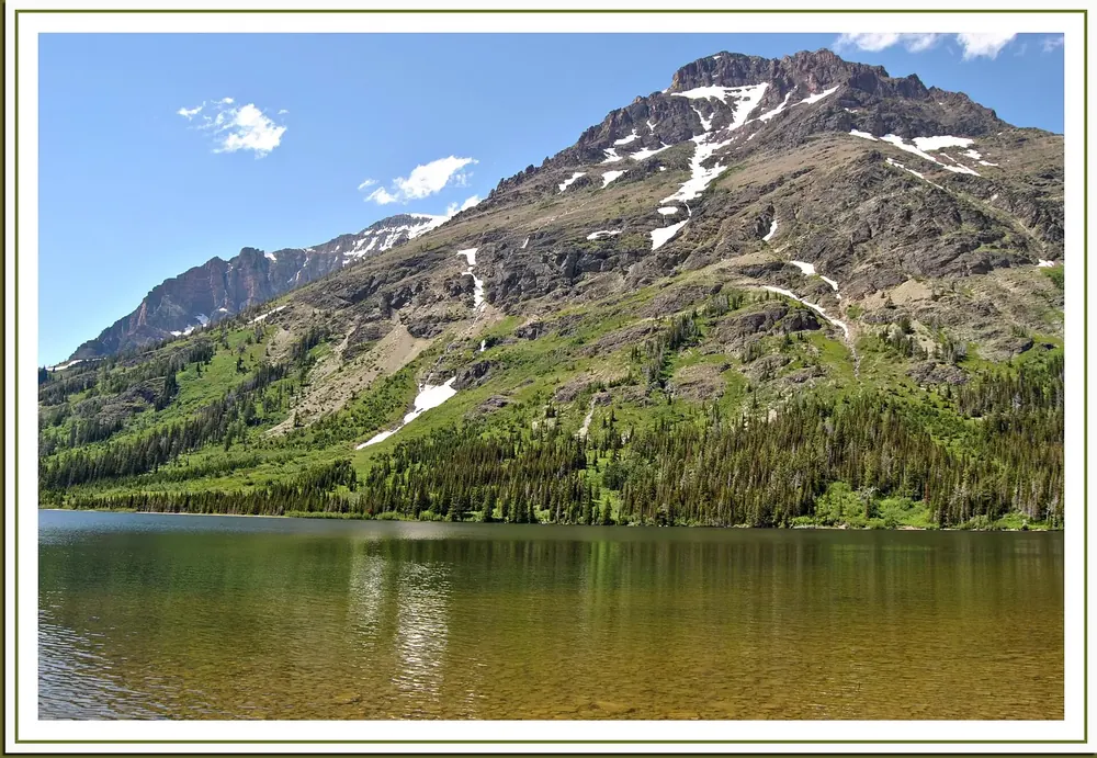 Two Medicine Lake: klarer Bergsee vor einem grünen Berghang mit Schneeresten unter blauem Himmel